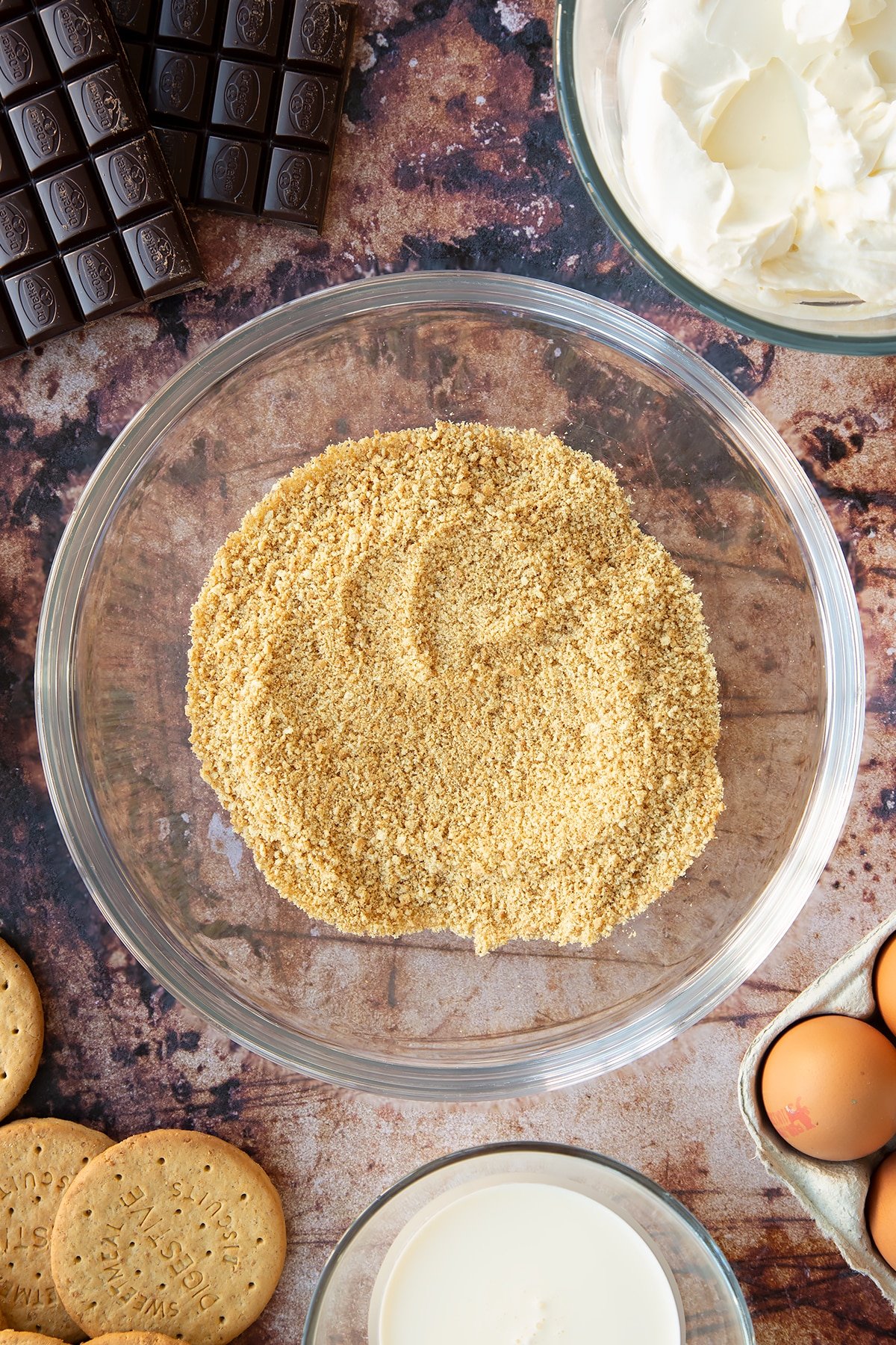 Crushed digestive biscuits in a glass bowl. Ingredients to make chocolate cheesecake cupcakes surround the bowl.