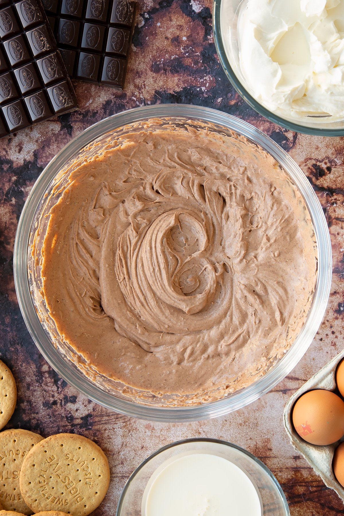 Chocolate cheesecake mix in a glass bowl. Ingredients to make chocolate cheesecake cupcakes surround the bowl.