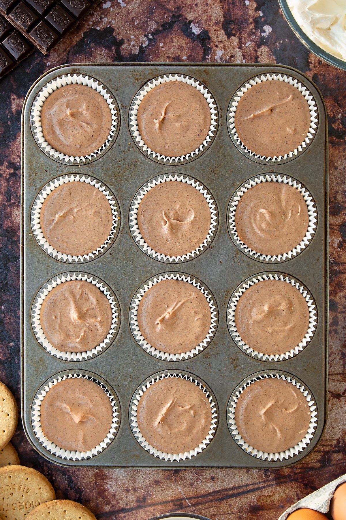 Chocolate cheesecake mix in cupcake cases in a muffin tray. Ingredients to make chocolate cheesecake cupcakes surround the tray.