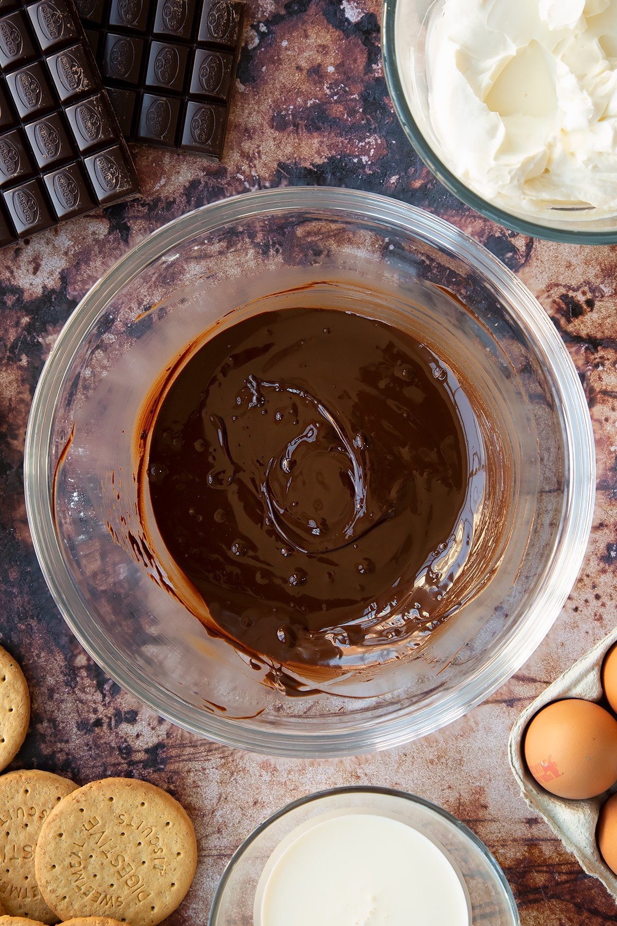 Melted dark chocolate in a glass bowl. Ingredients to make chocolate cheesecake cupcakes surround the bowl.