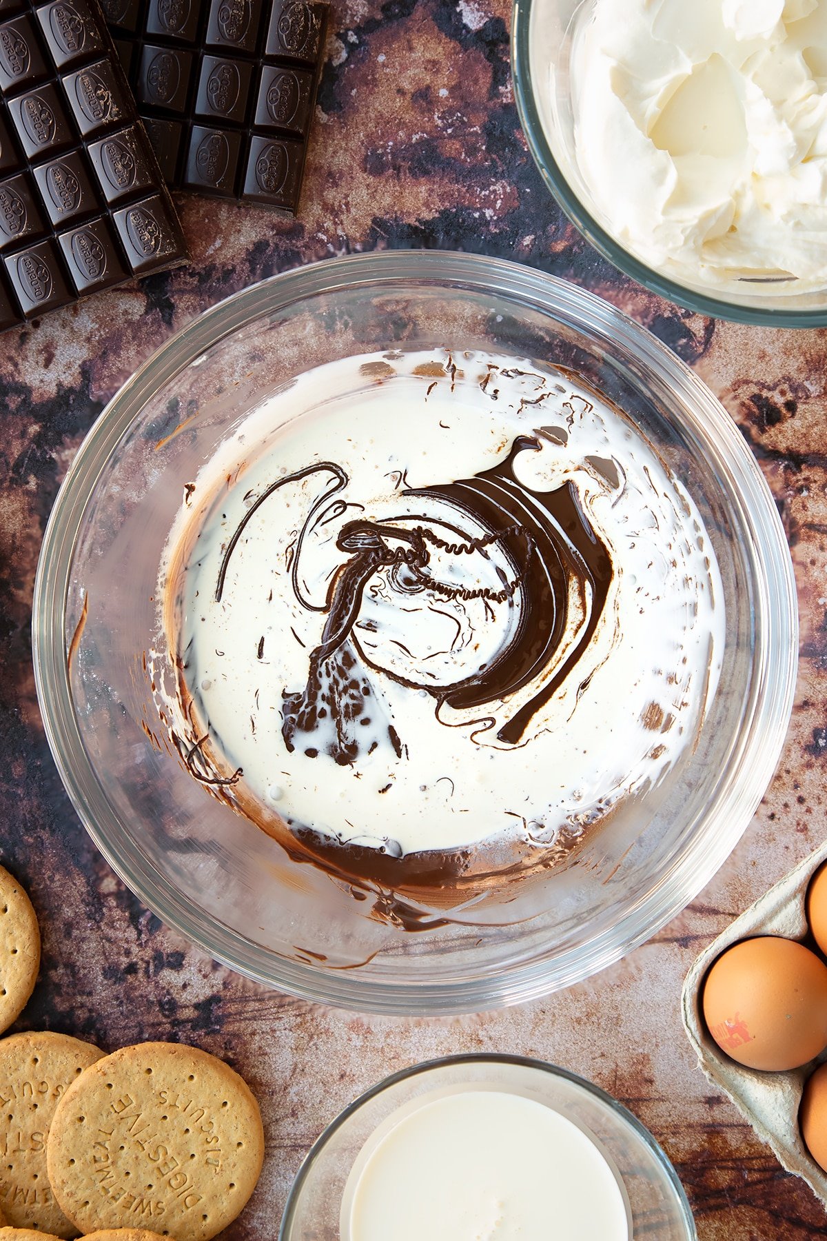 Melted dark chocolate and double cream in a glass bowl. Ingredients to make chocolate cheesecake cupcakes surround the bowl.