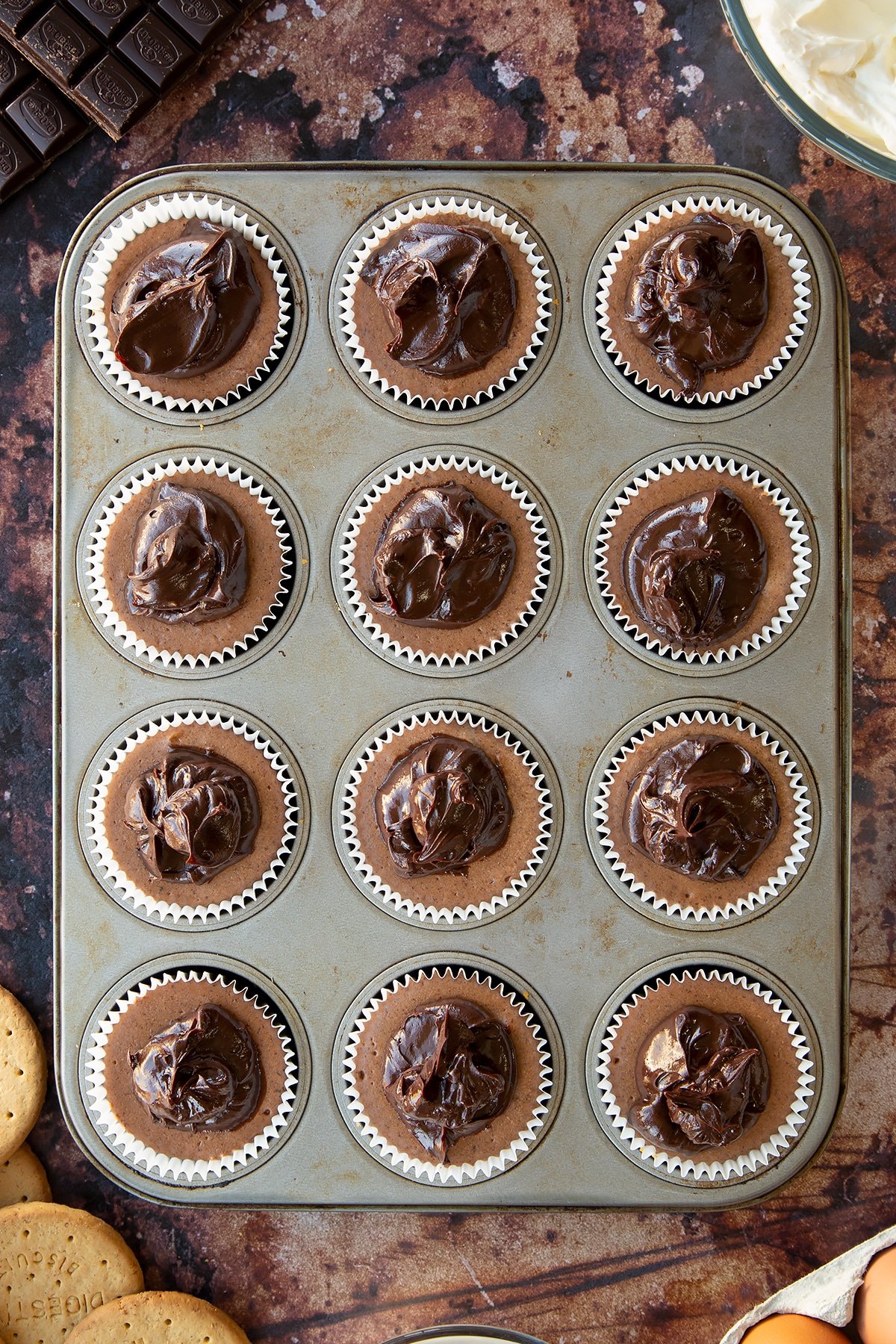 Chocolate cheesecake cupcakes, in a muffin tray. Each is topped with dollops of dark chocolate ganache.