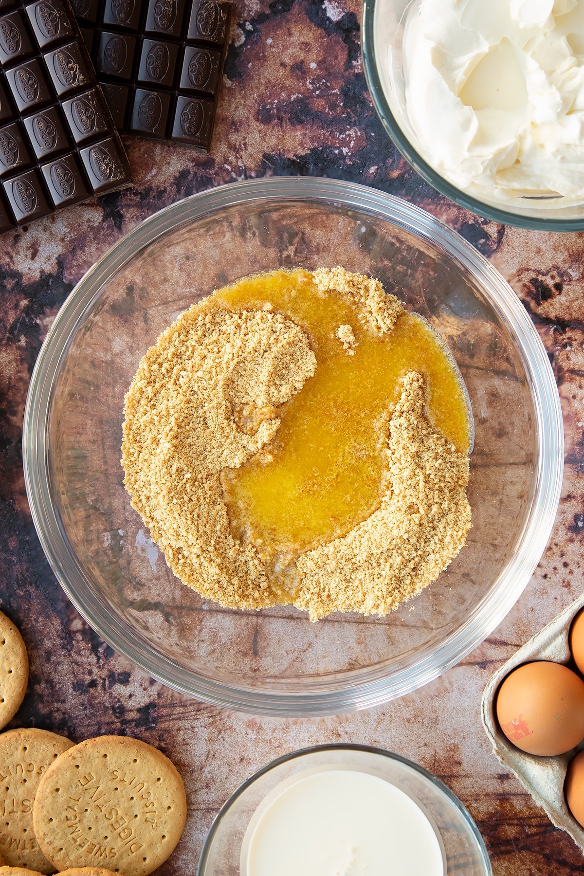 Crushed digestive biscuits and melted butter in a glass bowl. Ingredients to make chocolate cheesecake cupcakes surround the bowl.