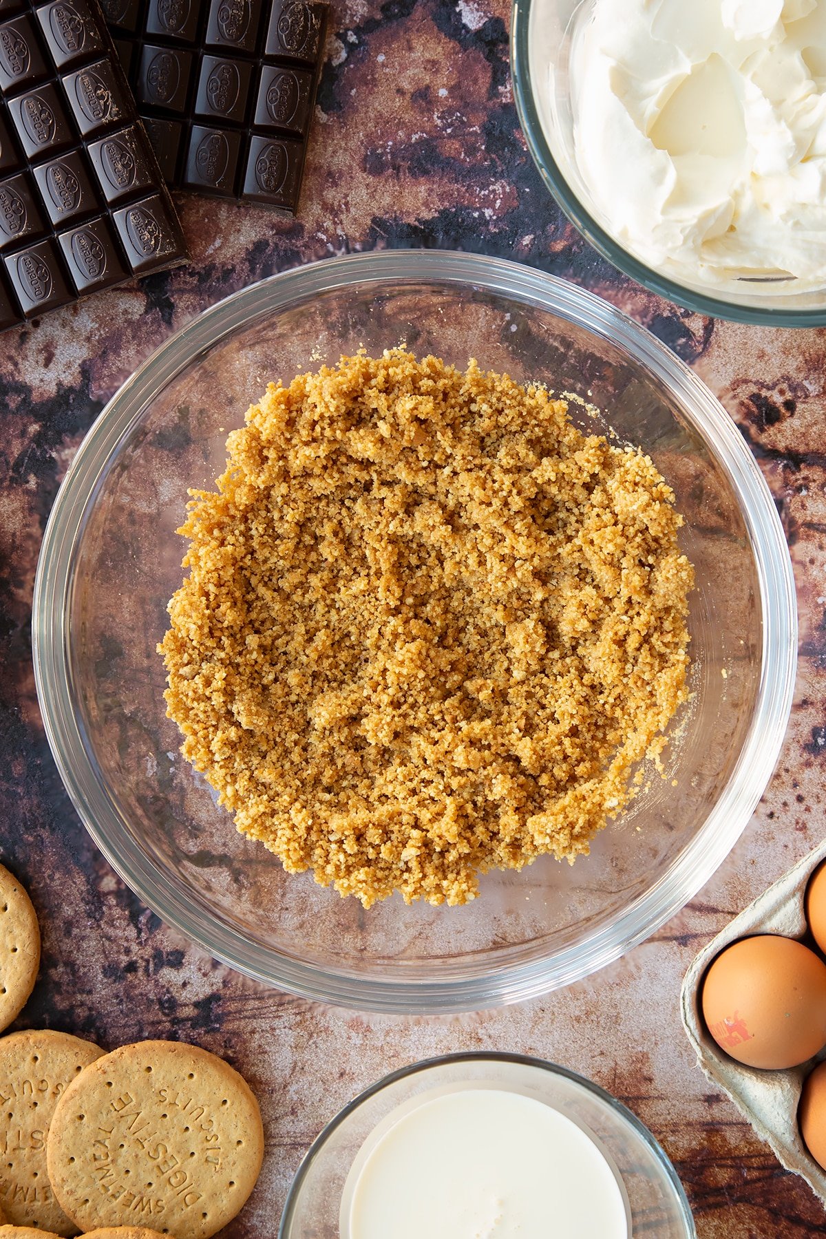 Crushed digestive biscuits mixed with melted butter in a glass bowl. Ingredients to make chocolate cheesecake cupcakes surround the bowl.