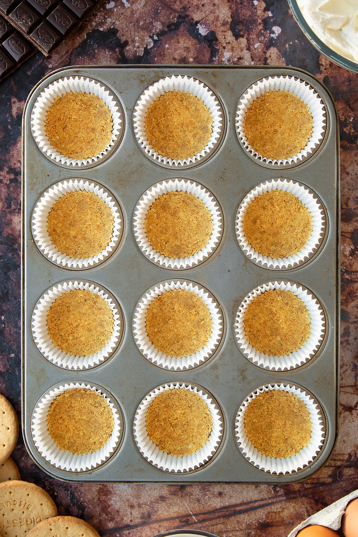 Crushed digestive biscuits mixed with melted butter pressed into cupcake cases in a muffin tray. Ingredients to make chocolate cheesecake cupcakes surround the tray.