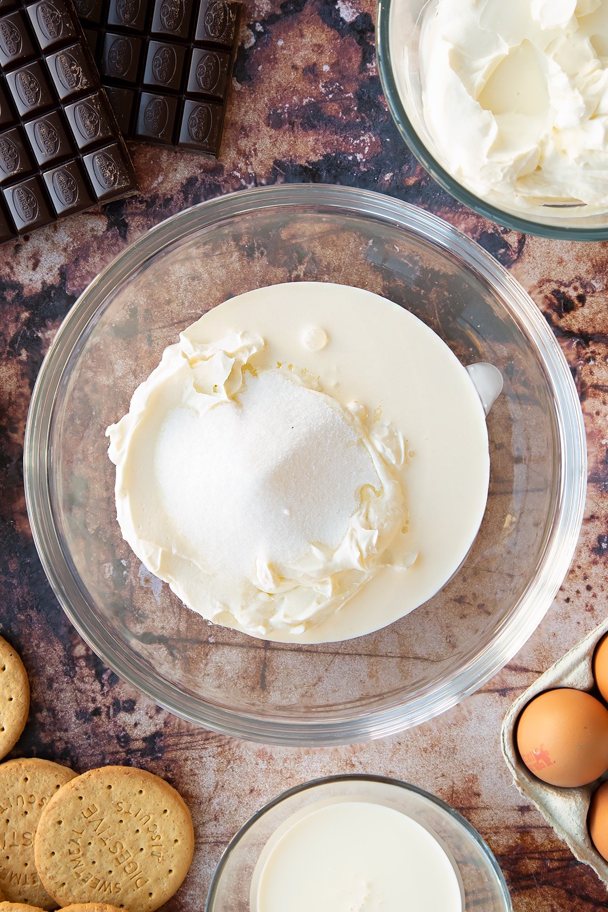 Cream cheese, sugar and cream in a glass bowl. Ingredients to make chocolate cheesecake cupcakes surround the bowl.
