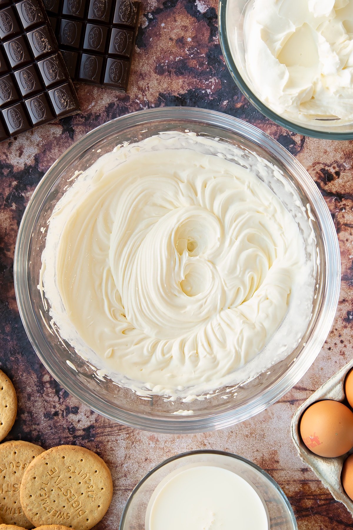 Cream cheese, sugar and cream whisked together in a glass bowl. Ingredients to make chocolate cheesecake cupcakes surround the bowl.