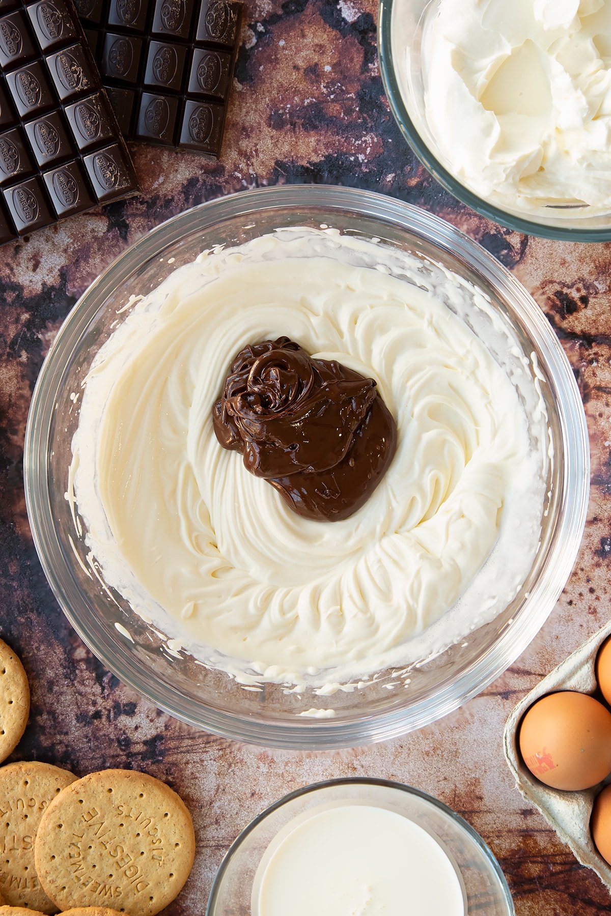 Cream cheese, sugar and cream whisked together in a glass bowl with dark chocolate on top. Ingredients to make chocolate cheesecake cupcakes surround the bowl.