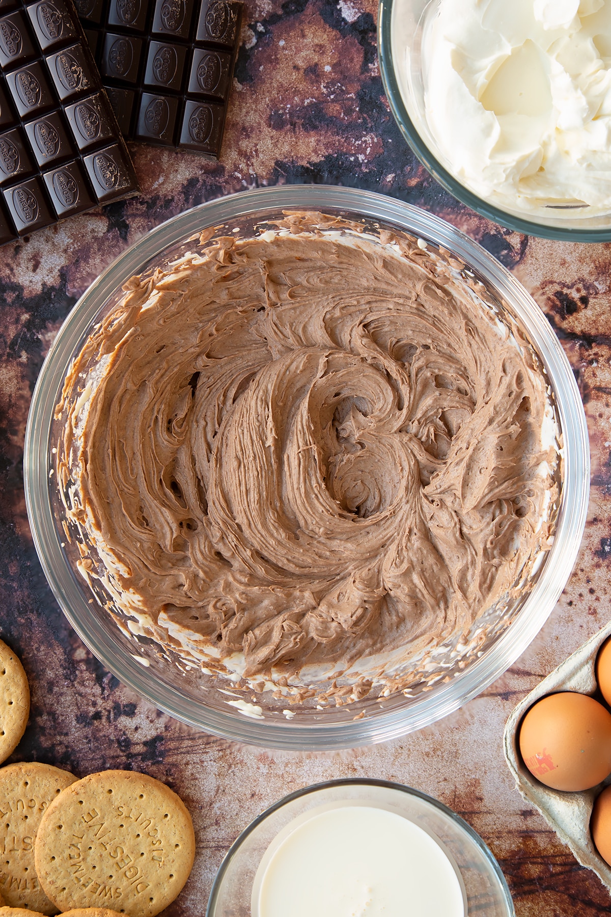 Cream cheese, sugar, cream and melted dark chocolate whisked together in a glass bowl. Ingredients to make chocolate cheesecake cupcakes surround the bowl.