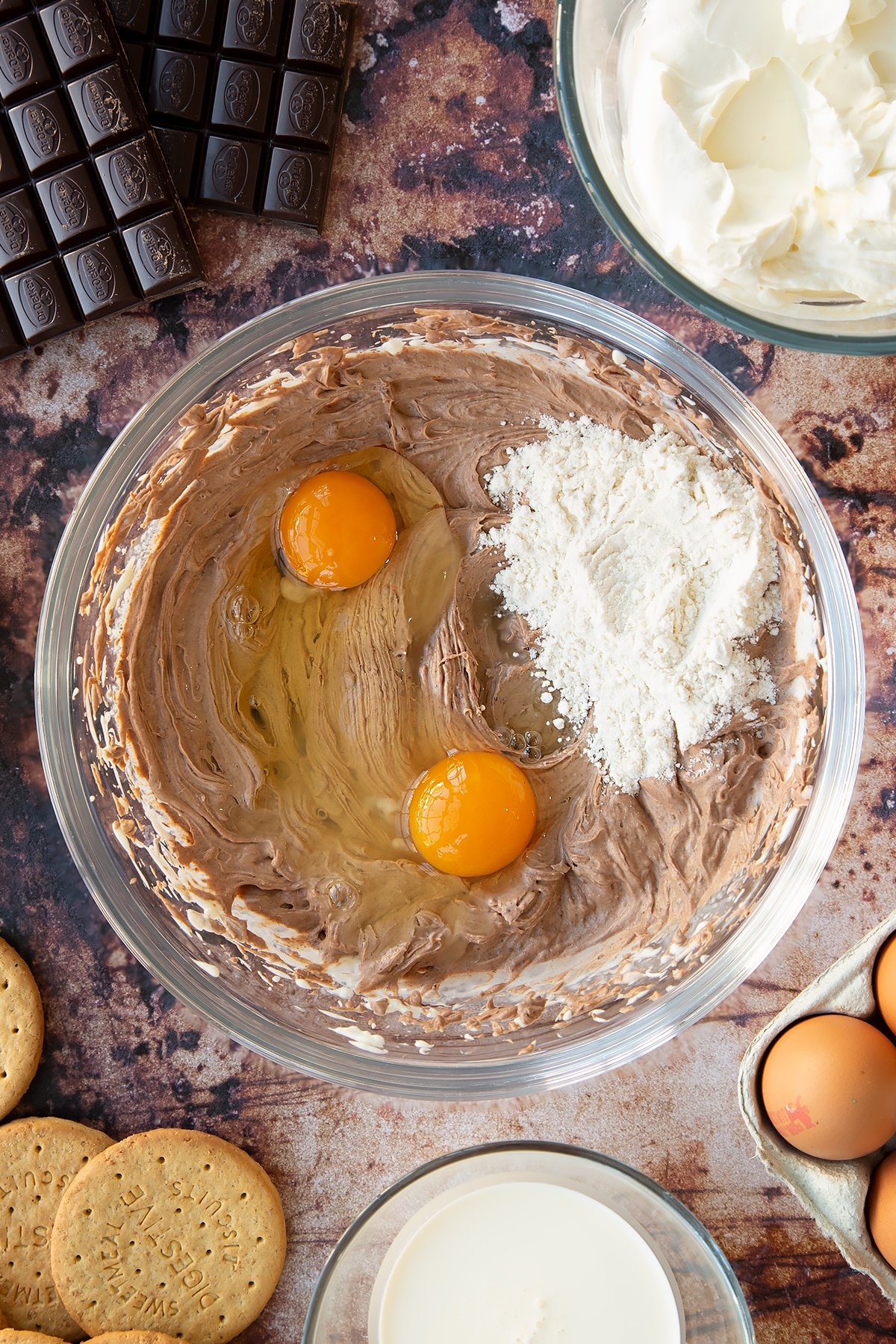 Cream cheese, sugar, cream and melted dark chocolate whisked together in a glass bowl with eggs and flour on top. Ingredients to make chocolate cheesecake cupcakes surround the bowl.