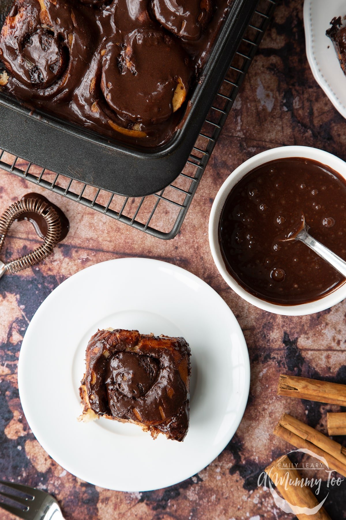 Overhead shot of chocolate cinnamon bun covered in chocolate sauce on a white plate with A Mummy Too logo in the corner