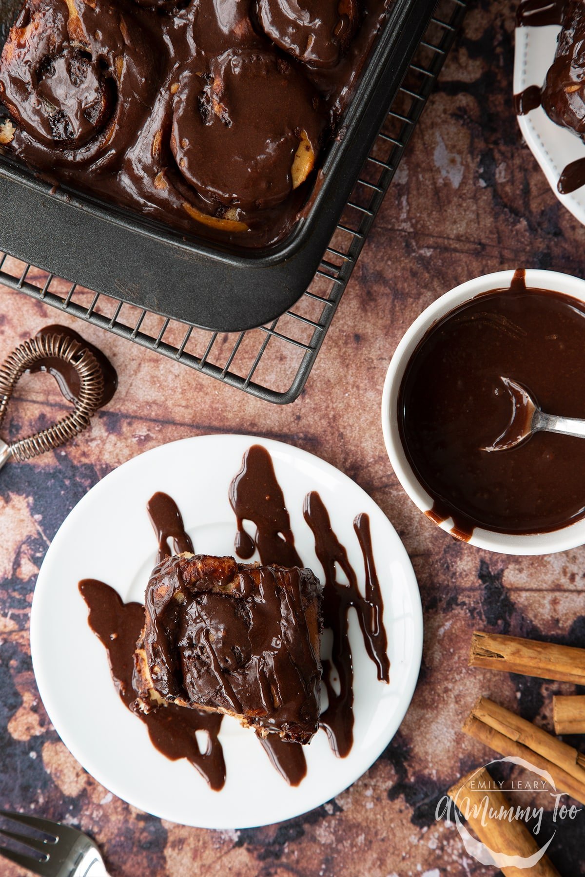 Overhead shot of a chocolate cinnamon bun drizzled with chocolate on a white plate with teh A Mummy Too logo in the corner