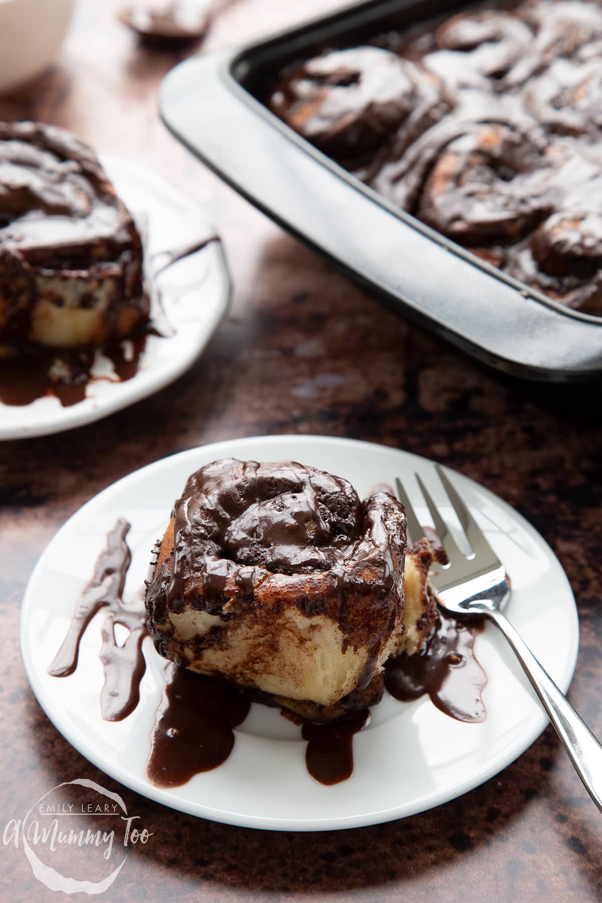 Front view of chocolate cinnamon bun with a fork on a white plate with the A Mummy Too logo in the corner