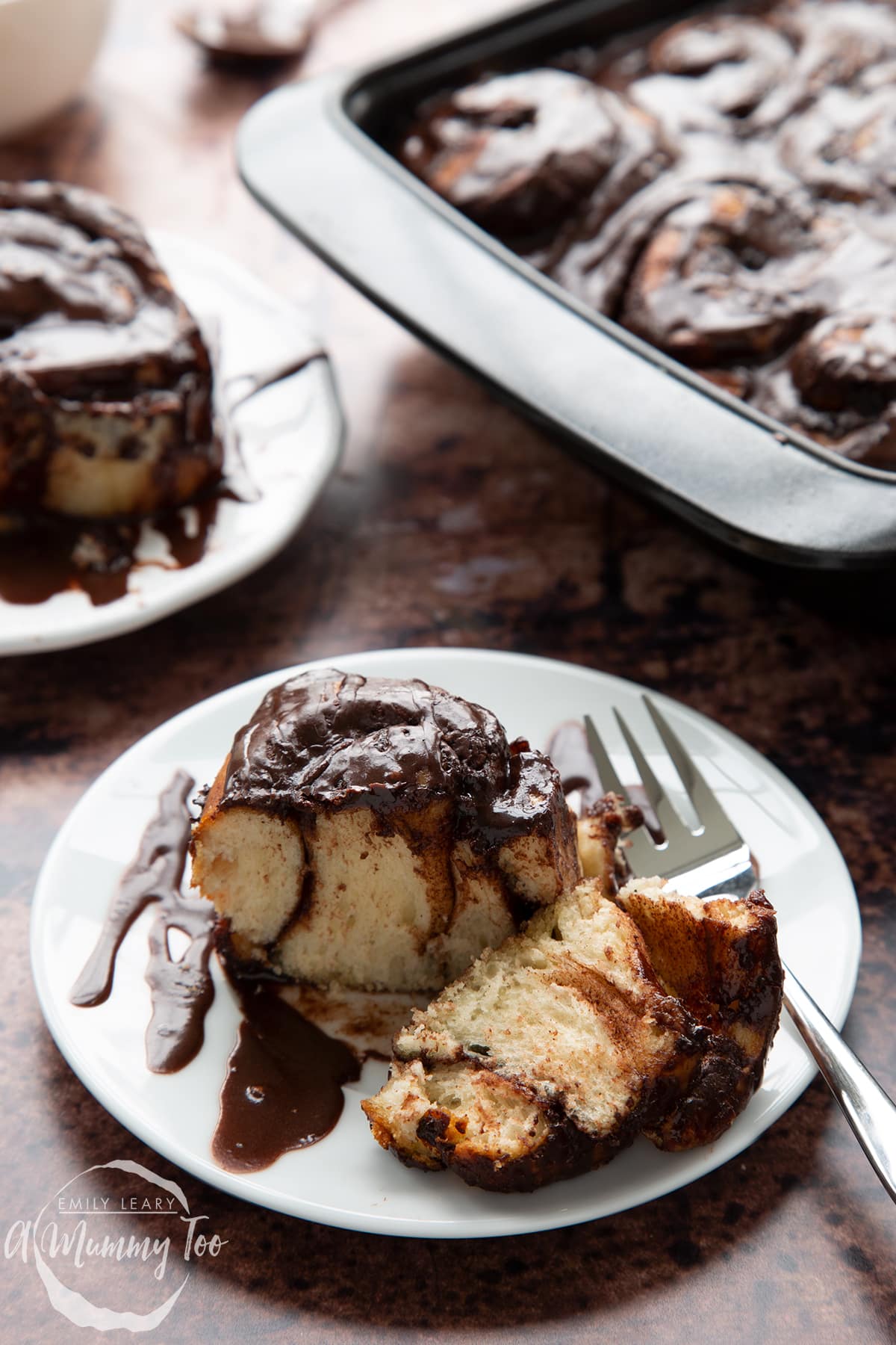 Front view of a chocolate cinnamon bun cut in half with a fork on a white plate with the A Mummy Too logo in the corner