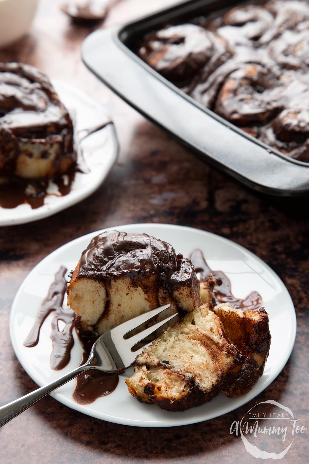 Front view of a chocolate cinnamon bun cut in half with a fork on a white plate with the A Mummy Too logo in the corner