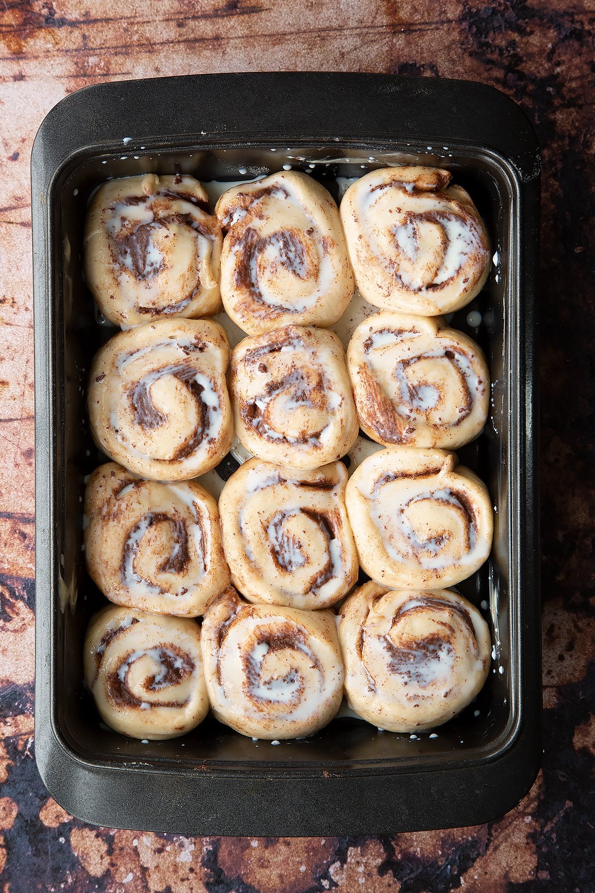 Overhead shot of Chocolate cinnamon bun dough rolled up into sections laid out in a deep baking tray covered with milk