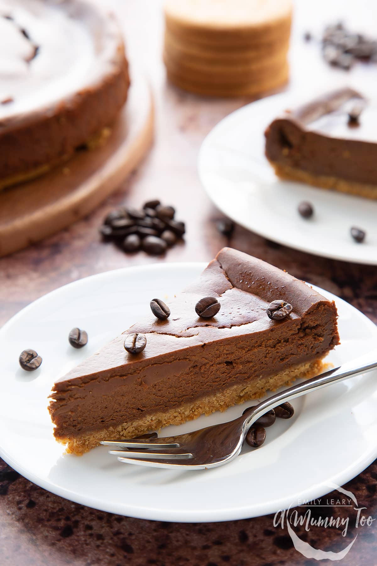 Front view shot of a piece of coffee cheesecake on a white plate topped with coffee beans with a fork on the side with the A Mummy Too logo in the corner