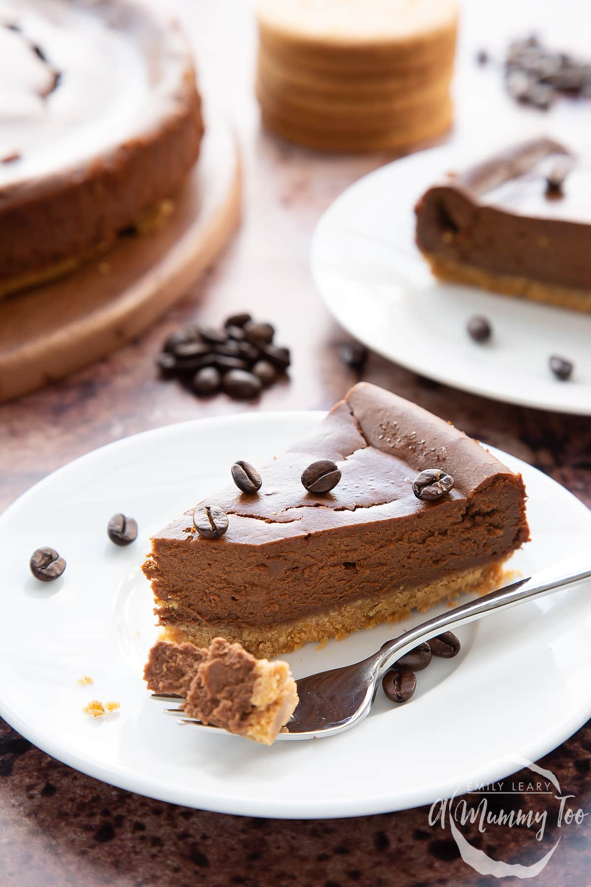 Front view shot of a piece of coffee cheesecake topped with coffee beans on a white plate with a fork with the tip of the cheesecake with the A Mummy Too logo in the corner