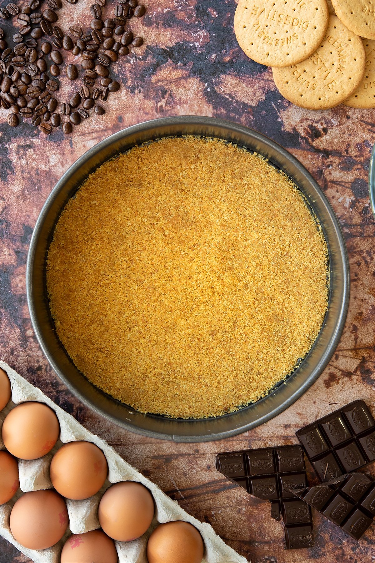 Overhead shot of butter and biscuit crumb mix pressed down flat in a large metal cake tin