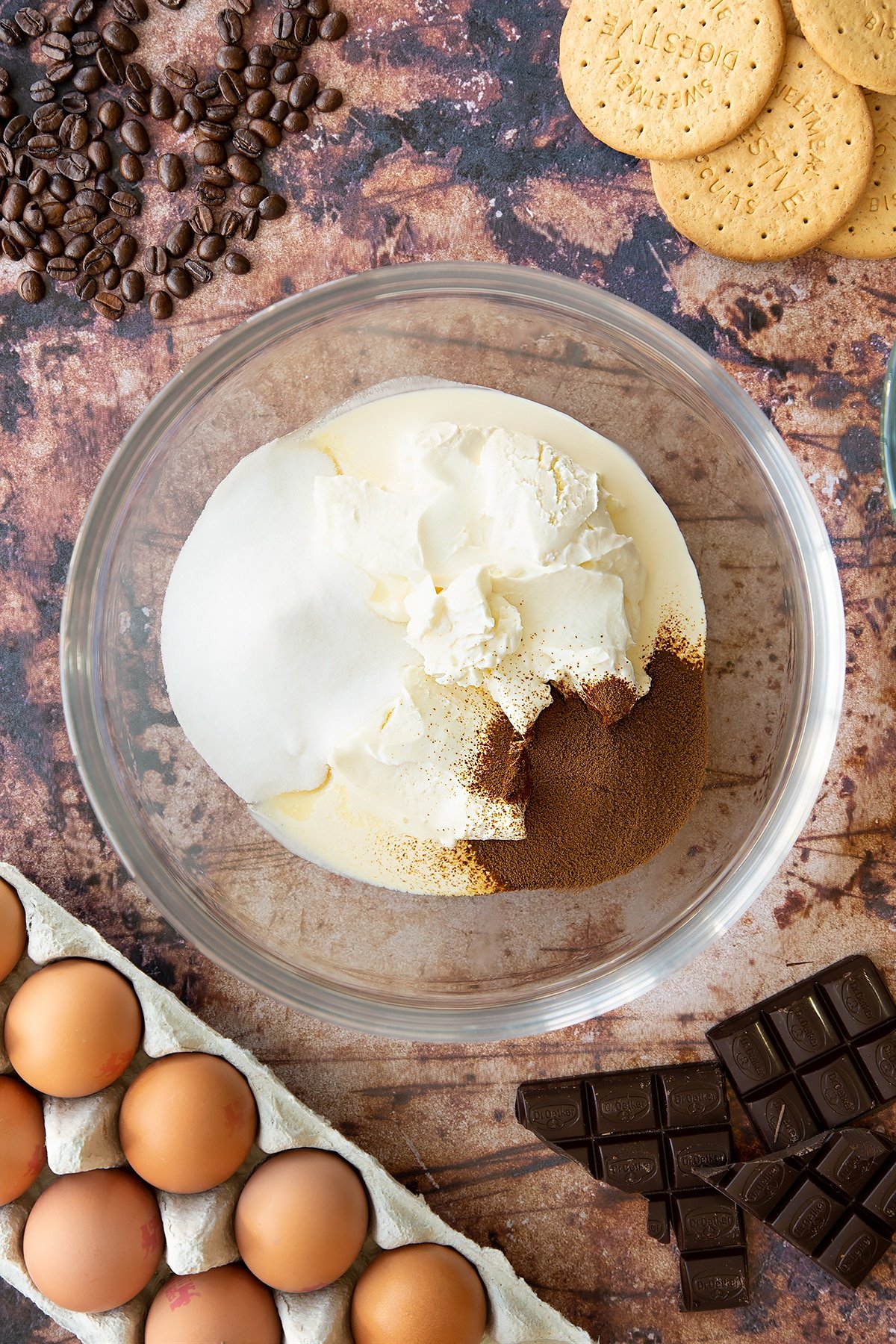 Overhead shot of cream cheese, cream, instant coffee and sugar in a large clear bowl