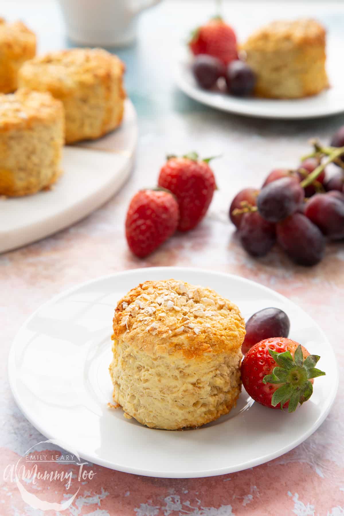 An oatmeal scone on a white plate with some fruit. More oatmeal scones sit on a board in the background.