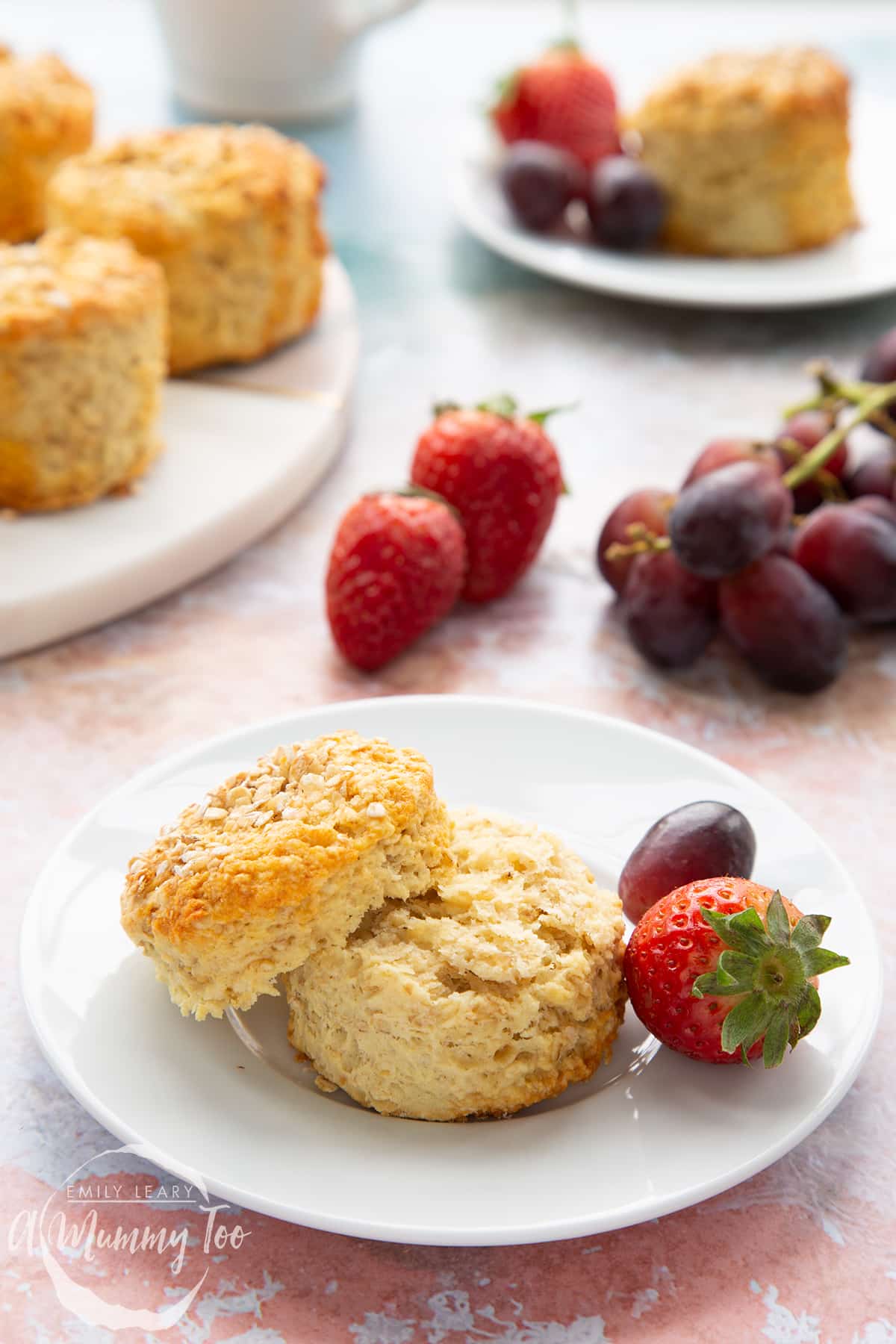 An oatmeal scone on a white plate with some fruit. The scone has been split. More oatmeal scones sit on a board in the background.