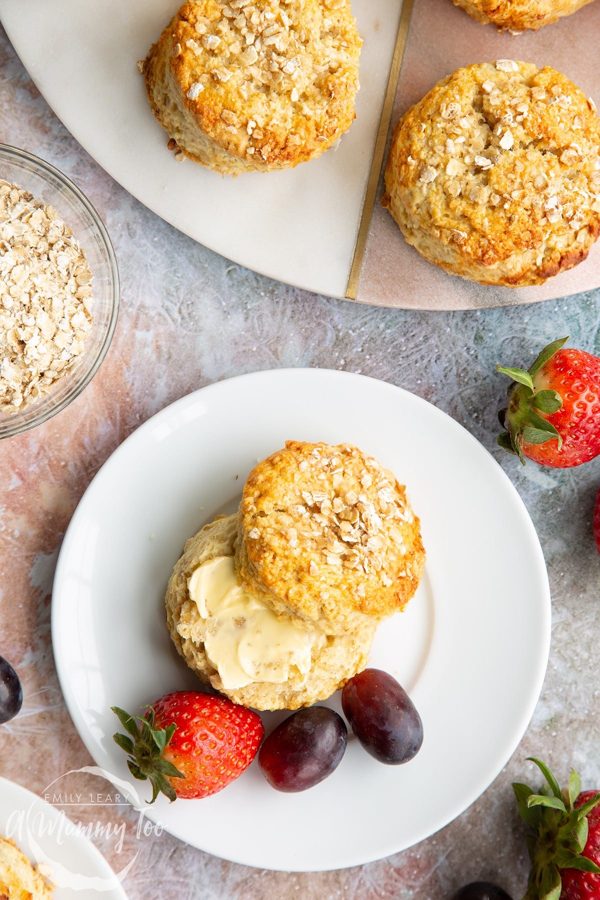 An oatmeal scone on a white plate with some fruit. The scone has been split and buttered. Shown from above.