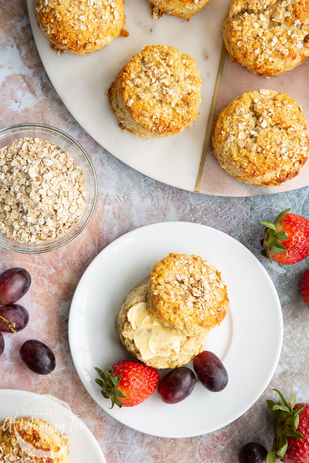 An oatmeal scone on a white plate with some fruit. Shown from above. There are more scones on a board to the edge of the frame.