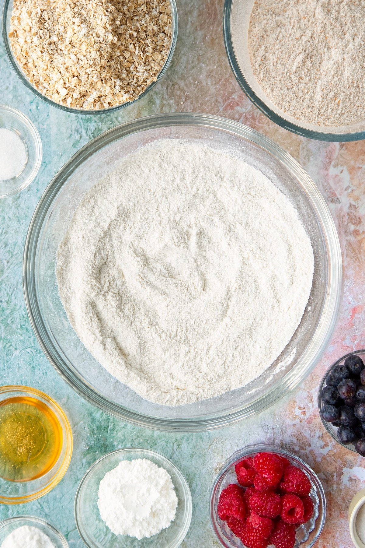 Self-raising flour, bicarbonate of soda and sugar combined in a glass mixing bowl. Ingredients to make oatmeal scones surround the bowl.