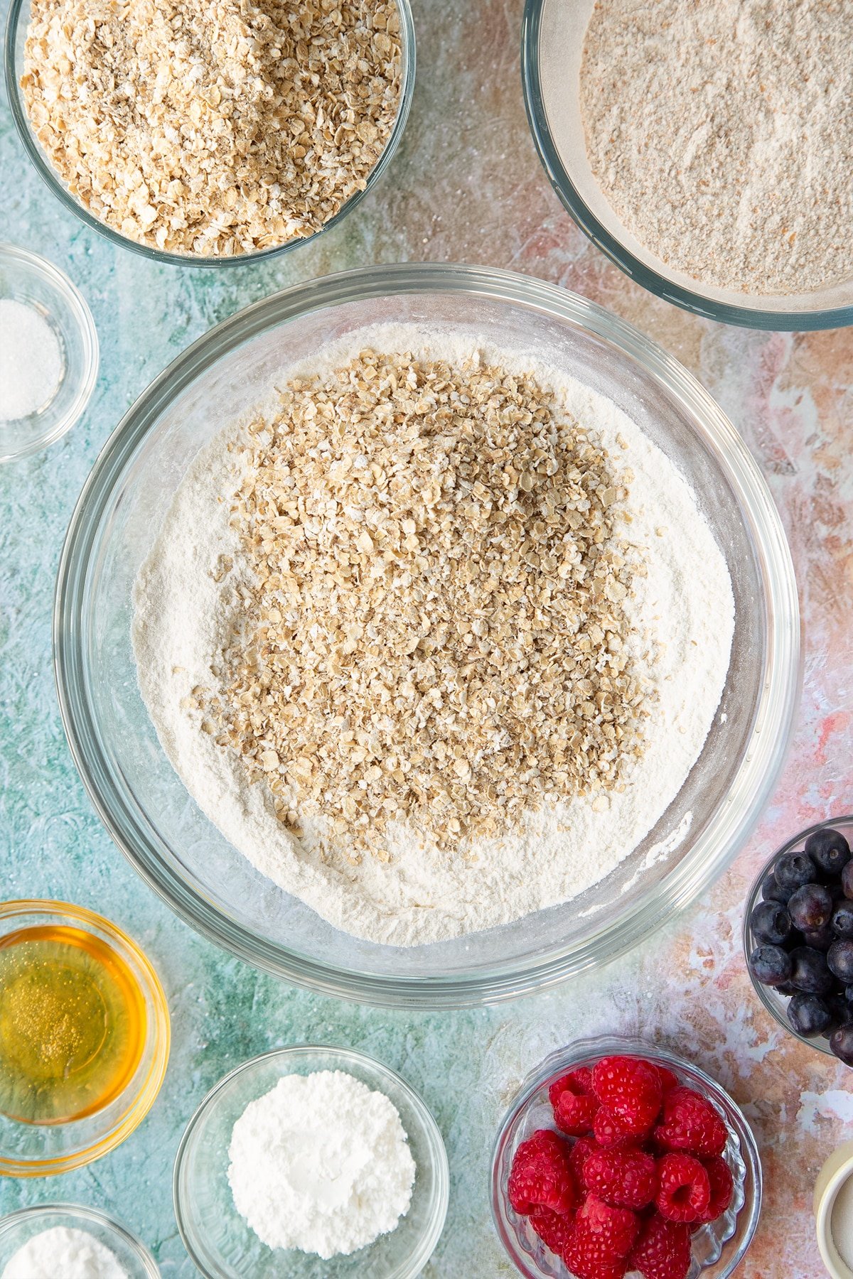 Self-raising flour, bicarbonate of soda and sugar combined in a glass mixing bowl with oats on top. Ingredients to make oatmeal scones surround the bowl.