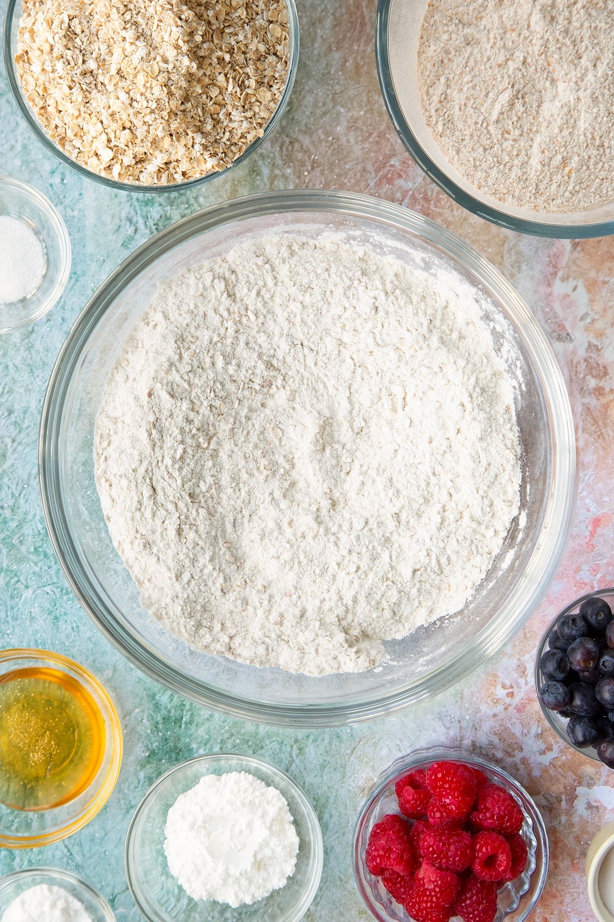 Self-raising flour, bicarbonate of soda, sugar and oats combined in a glass mixing bowl. Ingredients to make oatmeal scones surround the bowl.