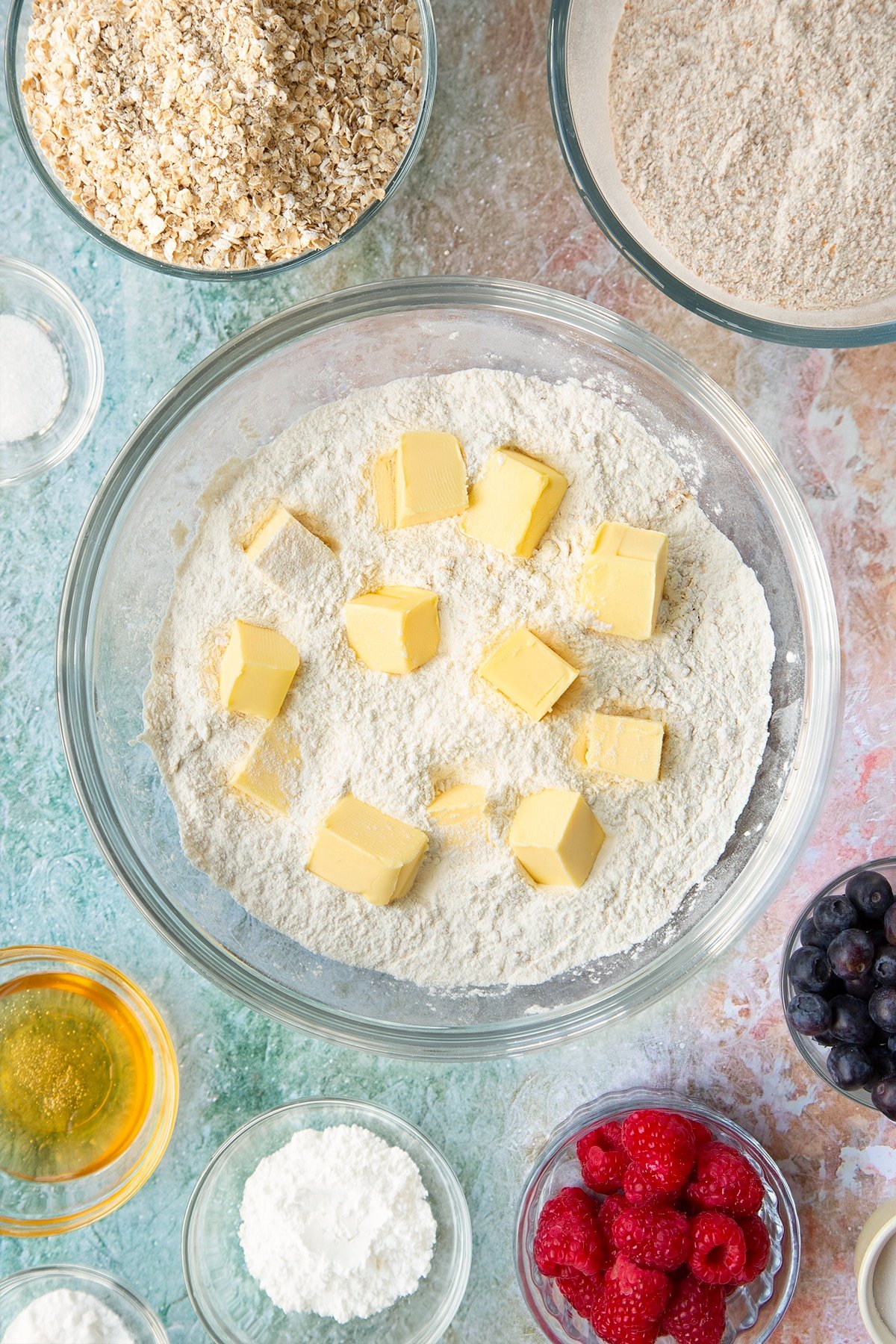 Self-raising flour, bicarbonate of soda, sugar and oats combined in a glass mixing bowl with cubed butter on top. Ingredients to make oatmeal scones surround the bowl.