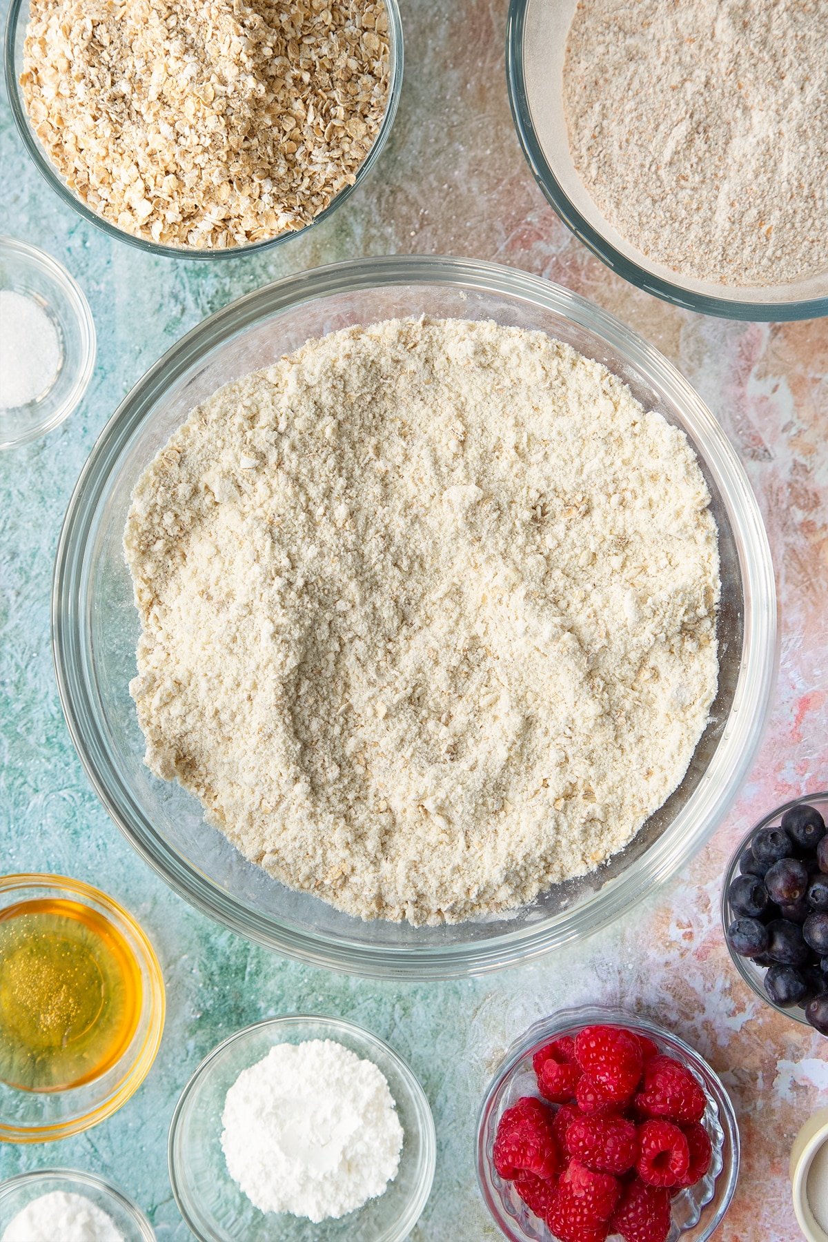 Butter rubbed into self-raising flour, bicarbonate of soda, sugar and oats in a glass mixing bowl. Ingredients to make oatmeal scones surround the bowl.