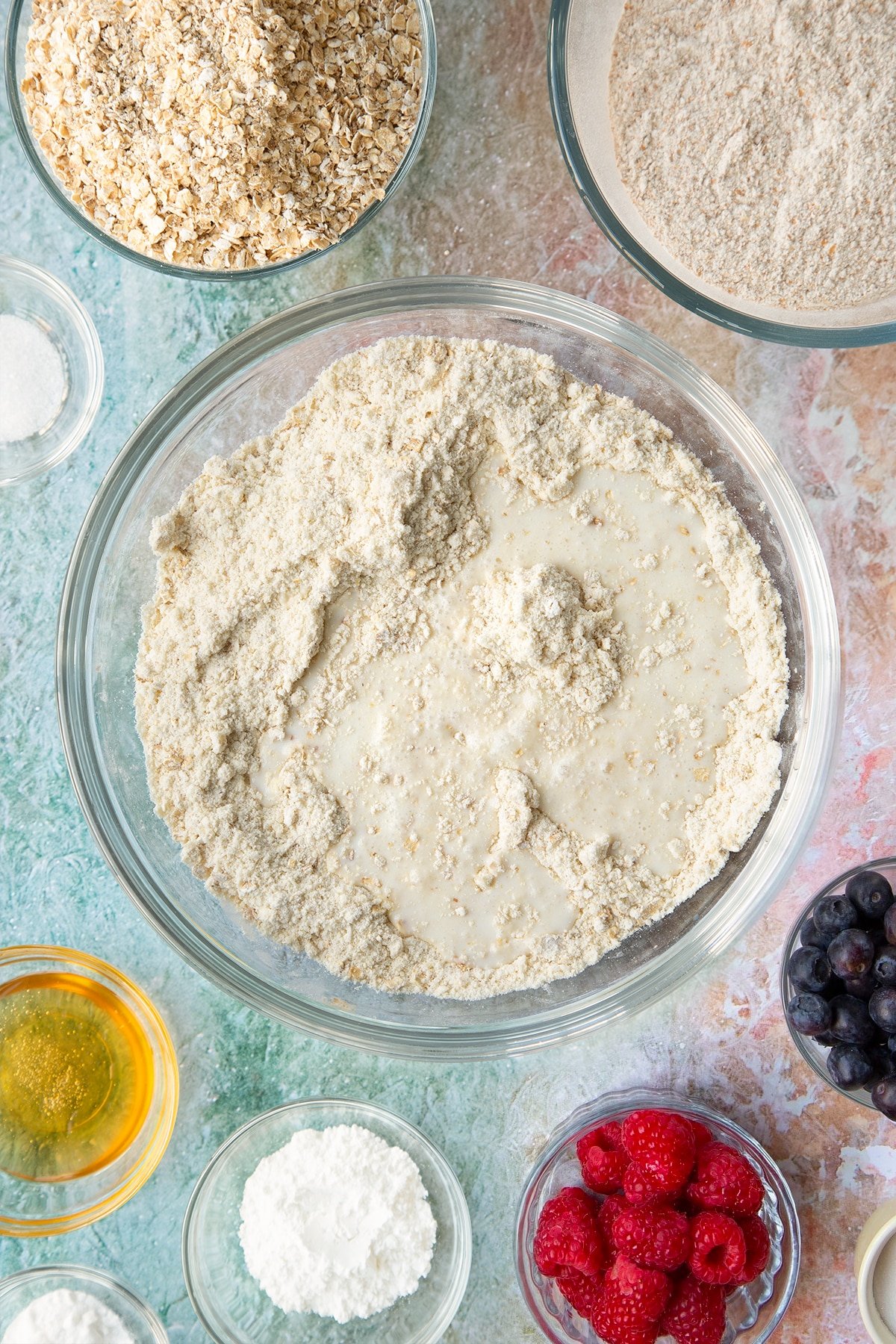 Butter rubbed into self-raising flour, bicarbonate of soda, sugar and oats in a glass mixing bowl with milk on top. Ingredients to make oatmeal scones surround the bowl.