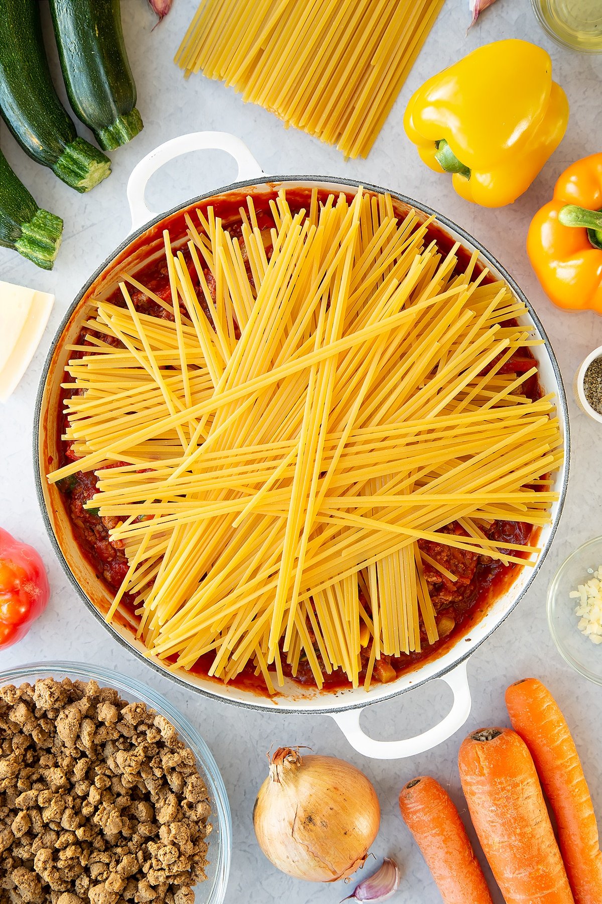 Veggie mince, garlic, onion, courgette, carrot, peppers and pasta sauce in a large pan with dried linguine on top. Ingredients to make one pot vegan bolognese surround the pan.