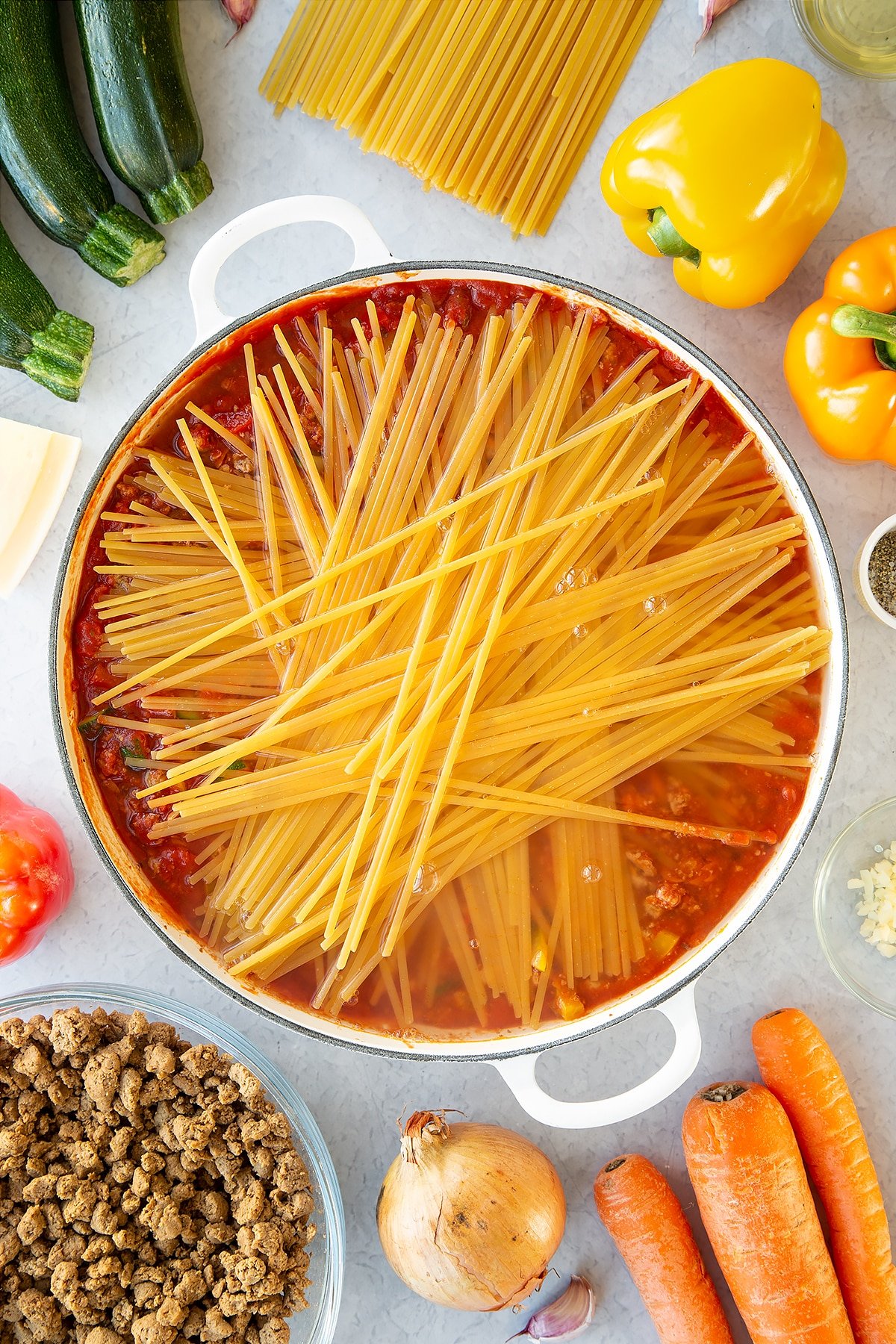 Veggie mince, garlic, onion, courgette, carrot, peppers and pasta sauce in a large pan with dried linguine on top, covered with boiling water. Ingredients to make one pot vegan bolognese surround the pan.