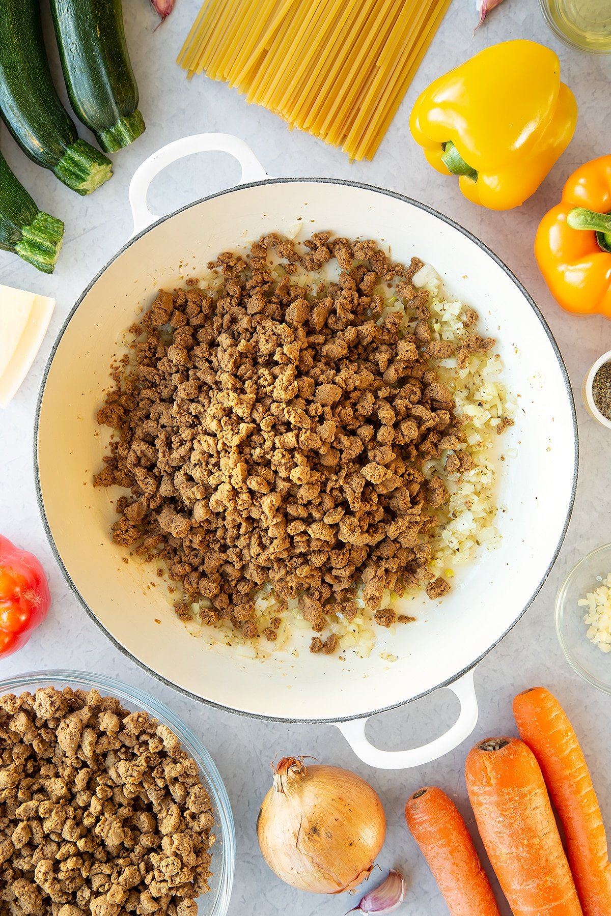 Seasoned garlic and onion sweated in a large pan with meat-free mince on top. Ingredients to make one pot vegan bolognese surround the pan.