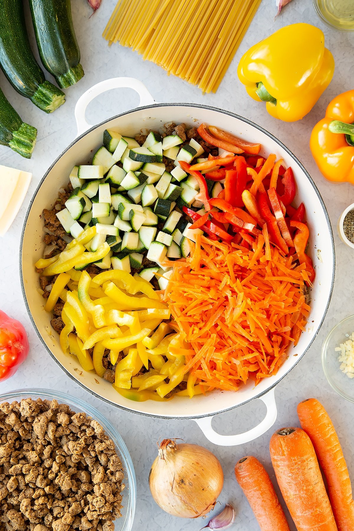 Veggie mince, garlic and onion sweated in a large pan with courgette, carrot and peppers on top. Ingredients to make one pot vegan bolognese surround the pan.