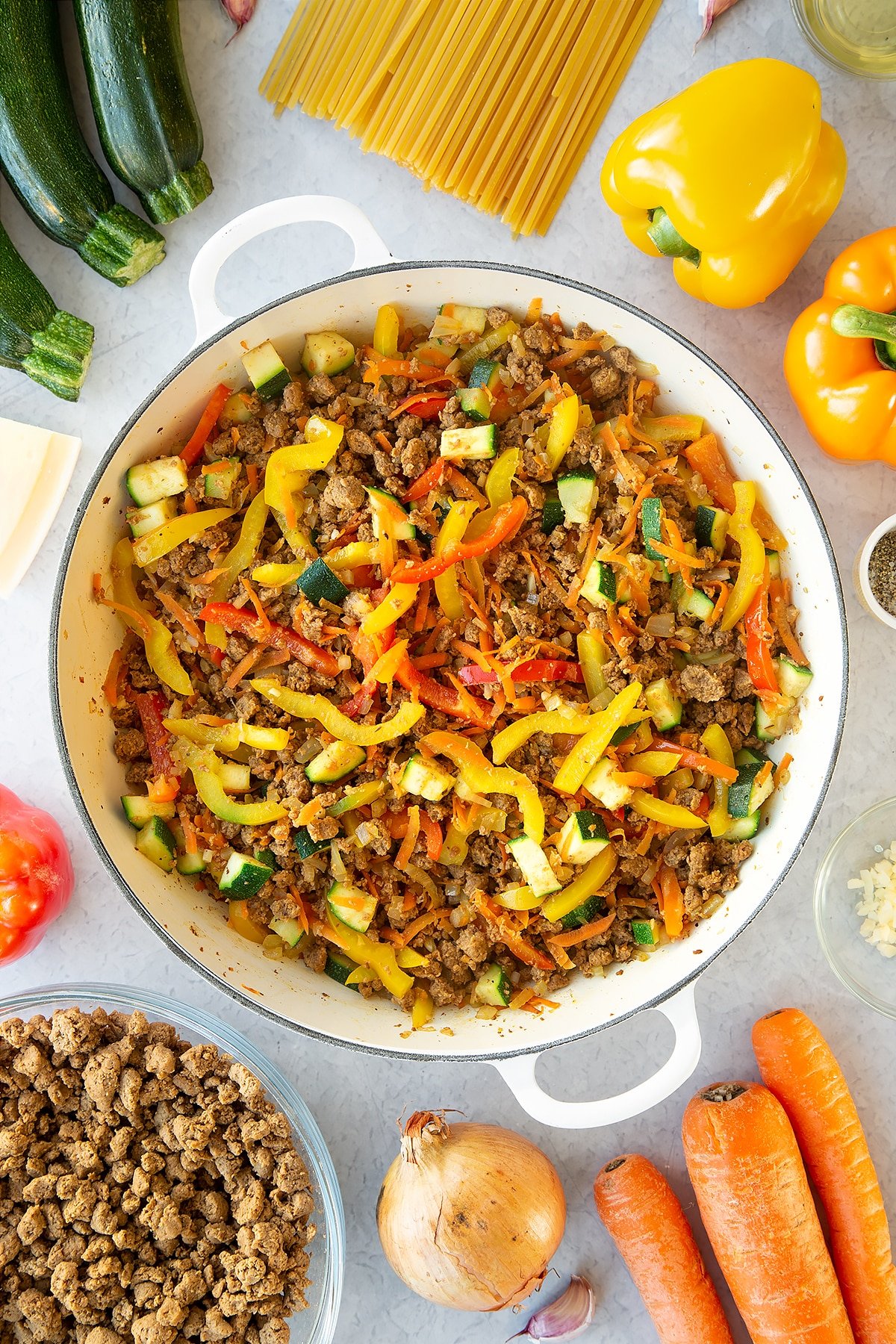 Veggie mince, garlic, onion, courgette, carrot and peppers sweated in a large pan. Ingredients to make one pot vegan bolognese surround the pan.