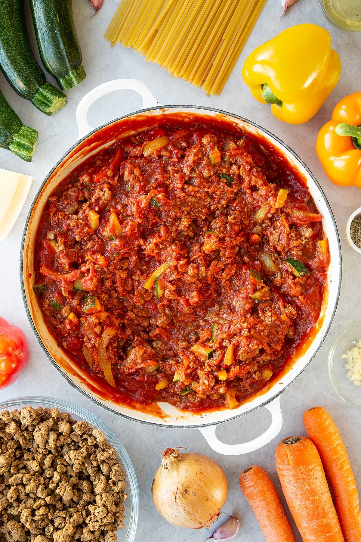 Veggie mince, garlic, onion, courgette, carrot, peppers and pasta sauce in a large pan. Ingredients to make one pot vegan bolognese surround the pan.