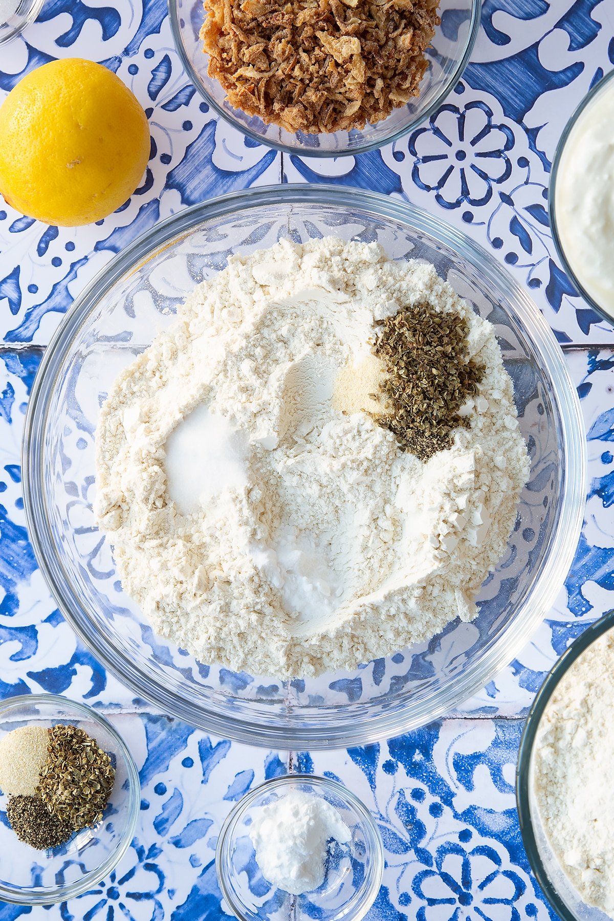 Flour, salt, bicarbonate of soda, garlic granules, oregano, salt and pepper in a mixing bowl. Ingredients to make onion soda bread surround the bowl.