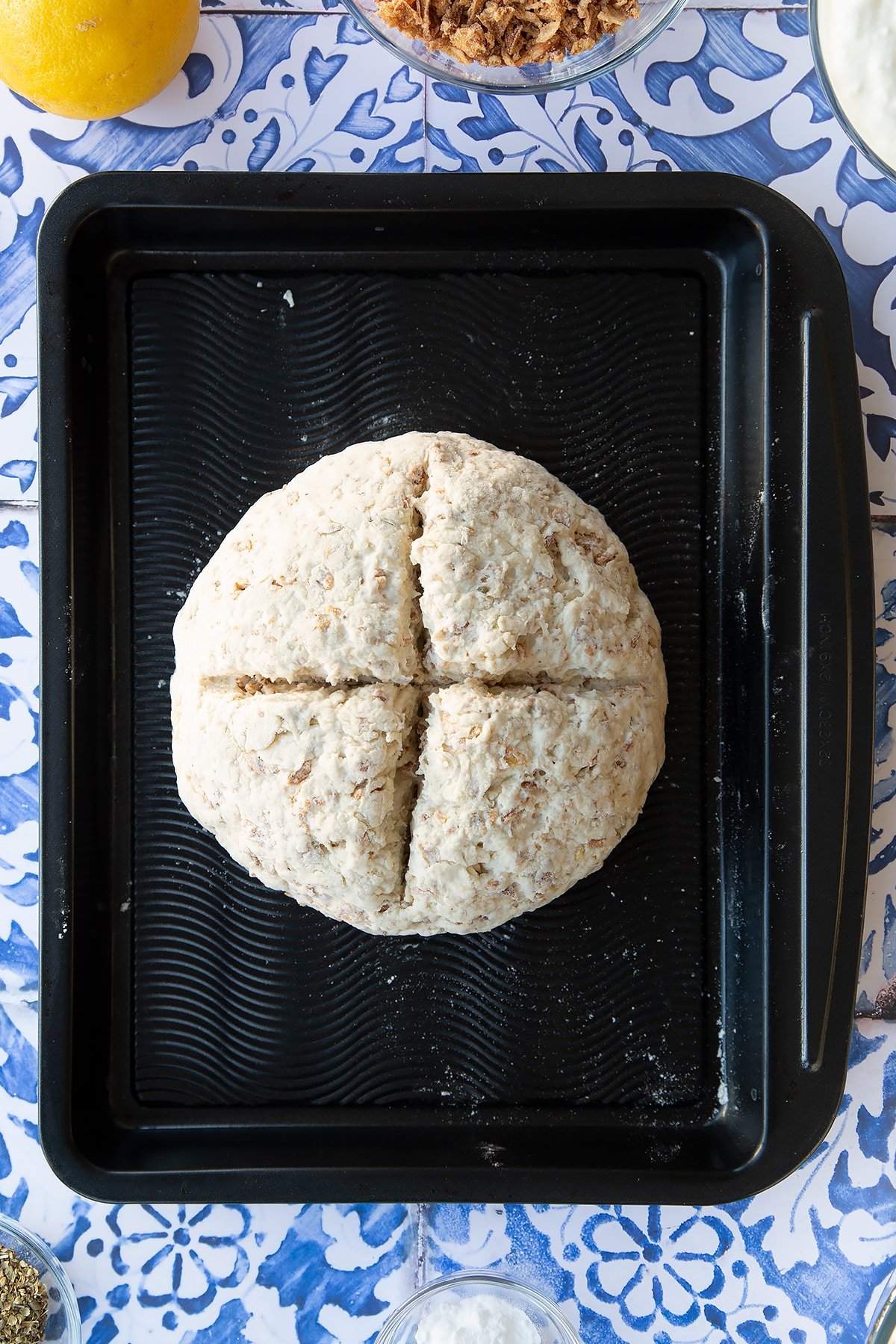 A ball of onion soda bread dough on a baking tray. A cross has been cut into the top. Ingredients to make onion soda bread surround the tray.