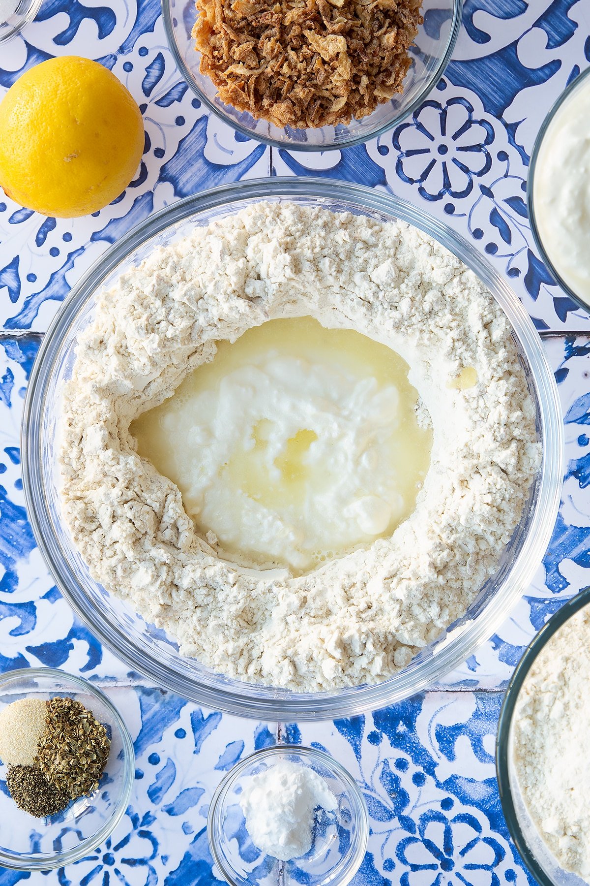 Flour, seasoning and crispy fried onions stirred together in a mixing bowl with a well in teh centre filled with yogurt and lemon juice. Ingredients to make onion soda bread surround the bowl.