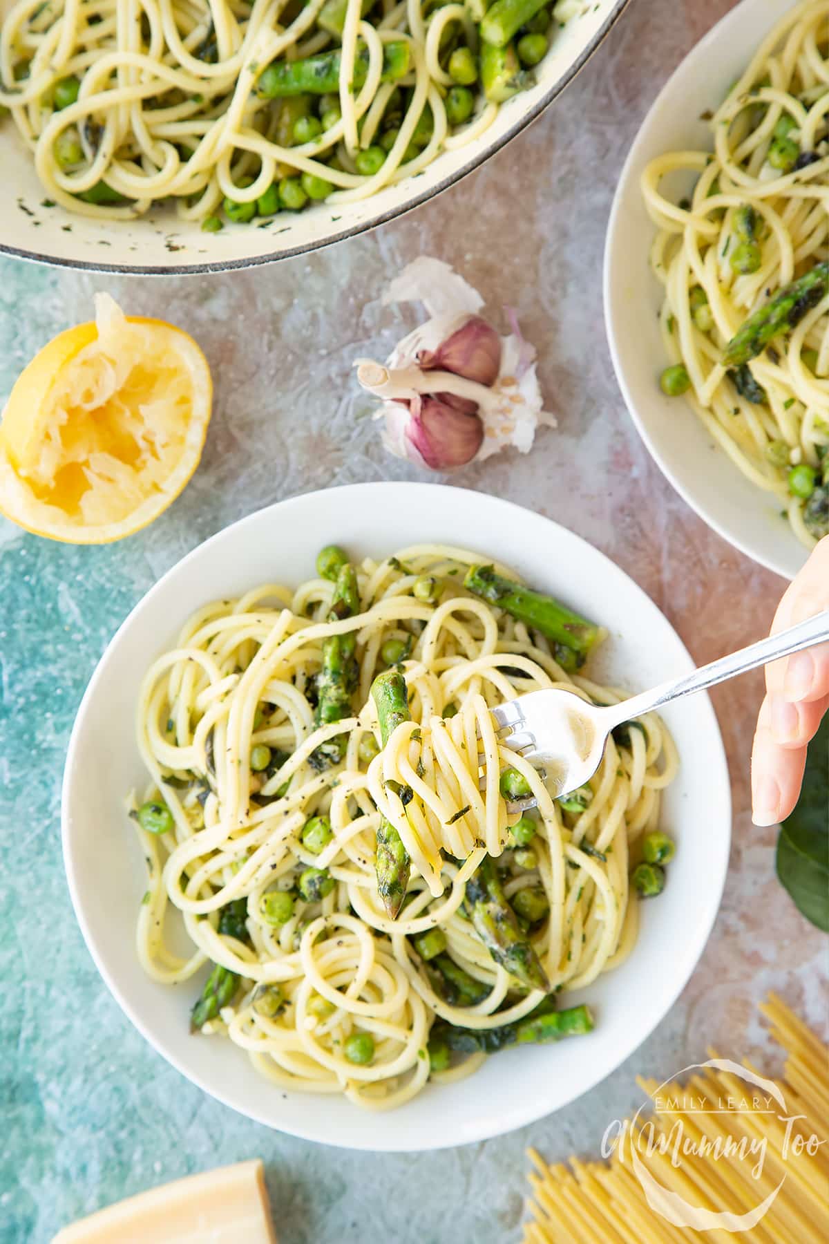 Summer spaghetti with spinach, basil, asparagus and peas, served in white bowls. Shown from above, a hand holds a fork with spaghetti twisted on it.
