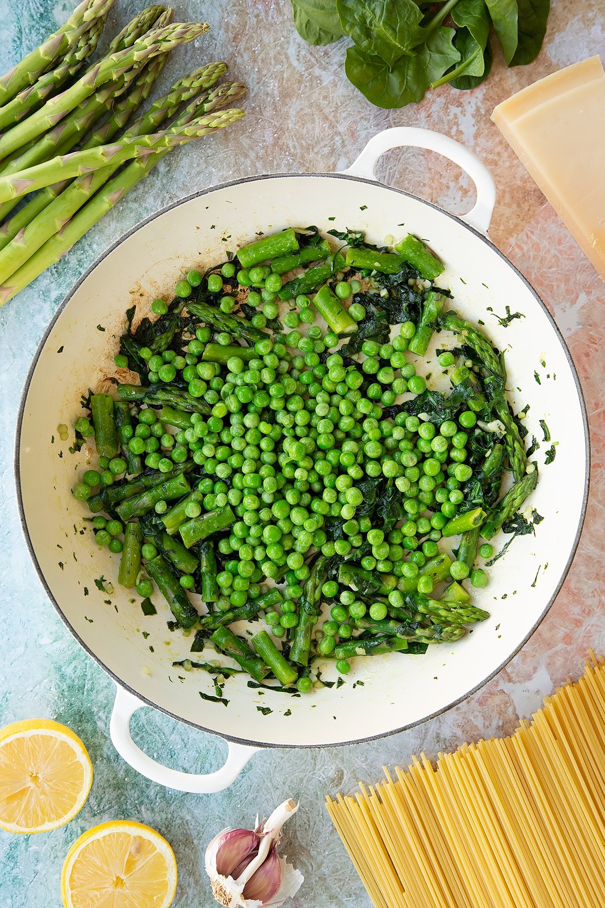 Lightly sautéed spinach, basil and asparagus with peas on top in a large, shallow saucepan. Ingredients to make summer spaghetti surround the pan.
