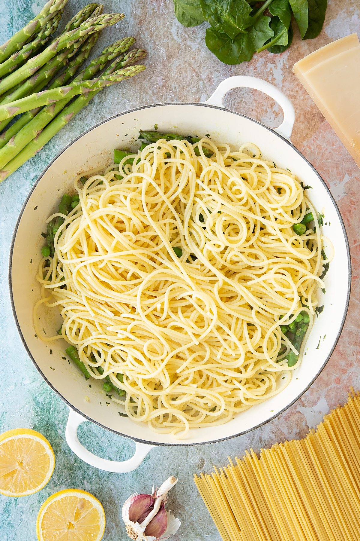 Lightly sautéed spinach, basil, asparagus and peas in a large, shallow saucepan with spaghetti on top. Ingredients to make summer spaghetti surround the pan.