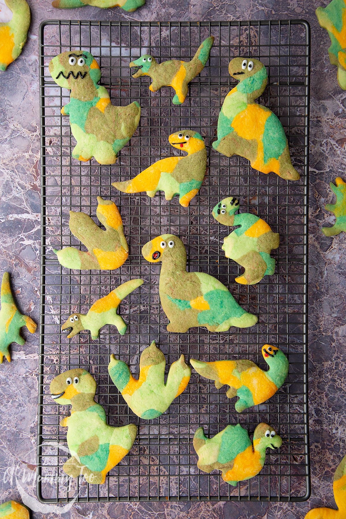 Overhead shot of baked multicoloured dinosaur cookie dough shapes with smiley faces on a wire rack