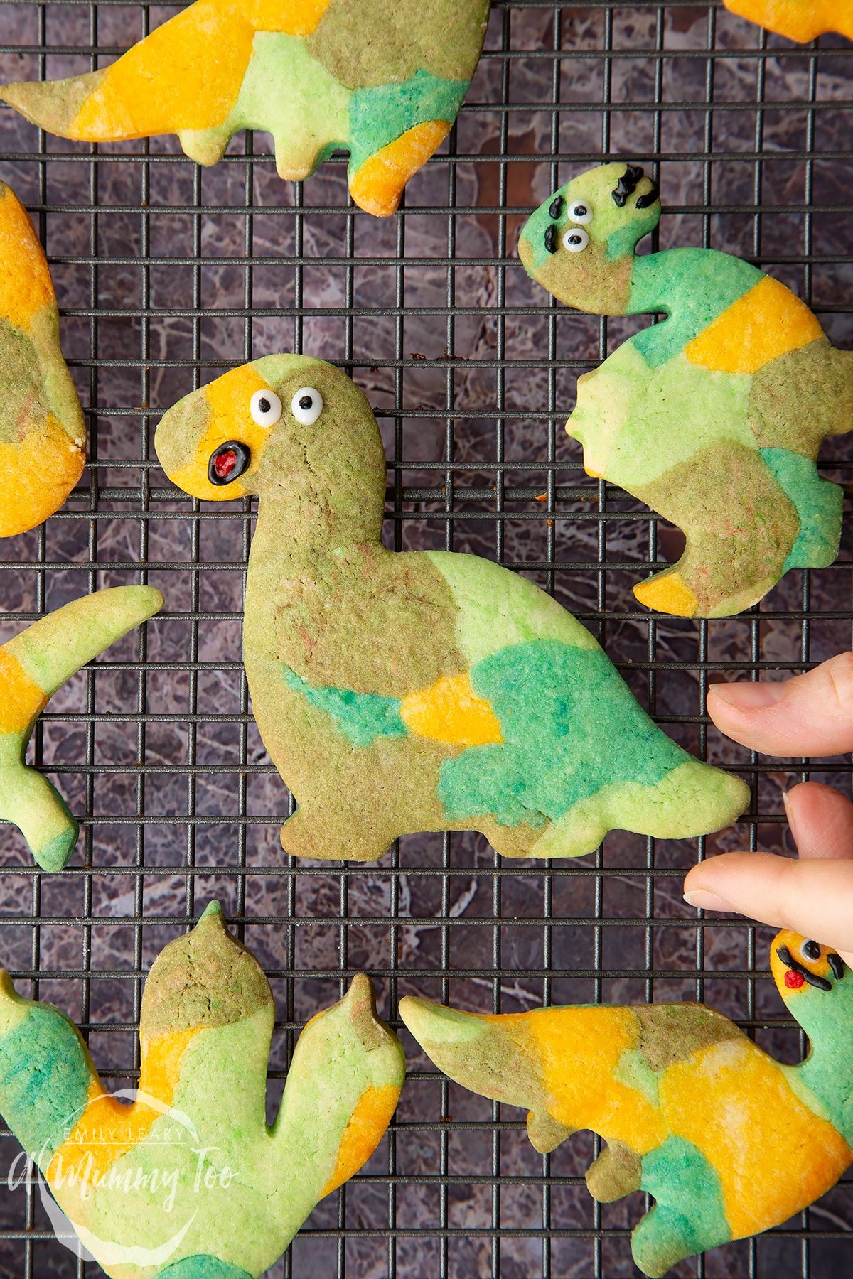 Overhead shot of baked multicoloured dinosaur cookie dough shapes with smiley faces on a wire rack