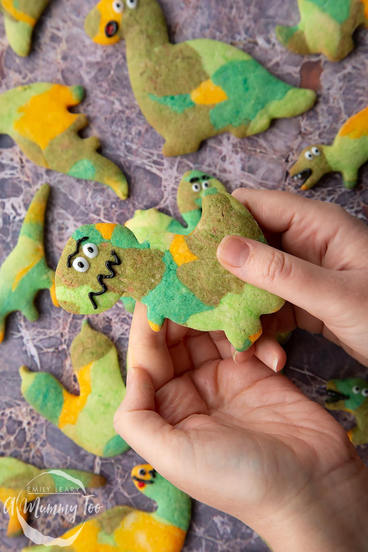 Overhead shot of a hand holding a green dinosaur cookie with the A Mummy Too logo in the corner