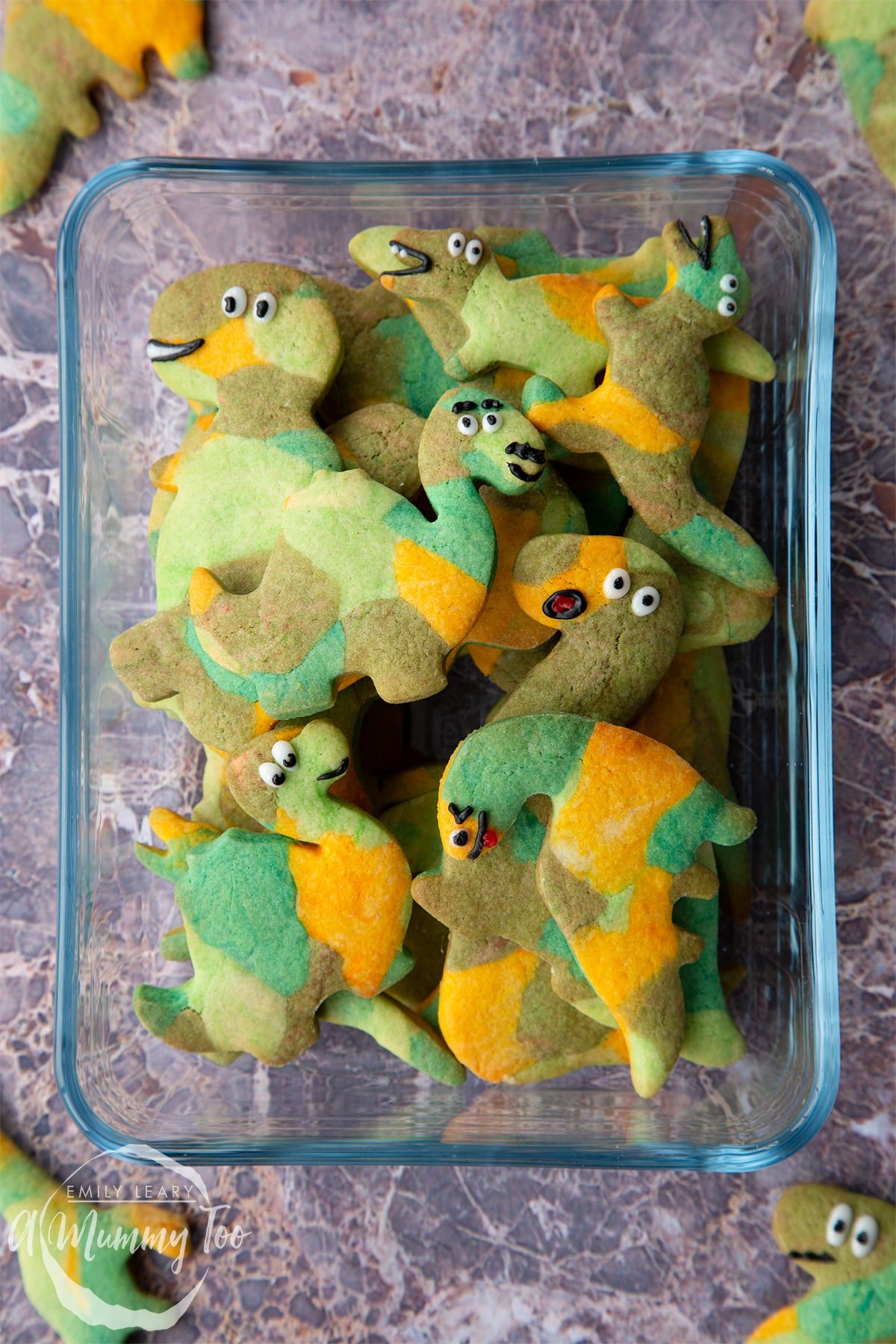 Overhead shot of dinosaur cookies stacked in a clear glass casserole dish with A Mummy Too logo in the corner